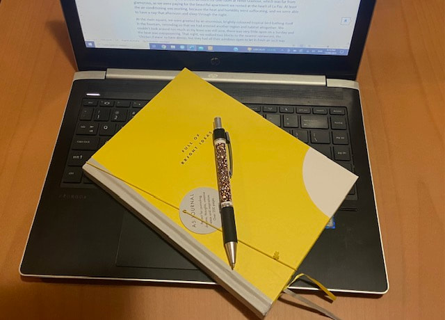 Image of a yellow A5 journal and a pen, sittin on a laptop's keyboard. The laptop is open and there's writing on the screen